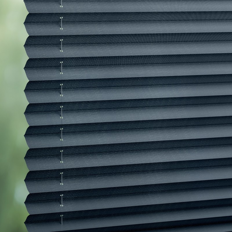 Dark gray pleated cellular blinds, stacked closed with visible cords and staples, blocking light; filtering soft daylight from a blurred green outdoor view through a window, creating diffuse interior lighting.