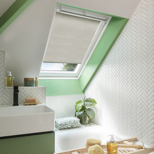 White pleated skylight blind, lowered halfway across a tilted window, filtering soft daylight; set in a green‑trimmed attic bathroom with white herringbone tiles, sink, tub, and a potted plant.