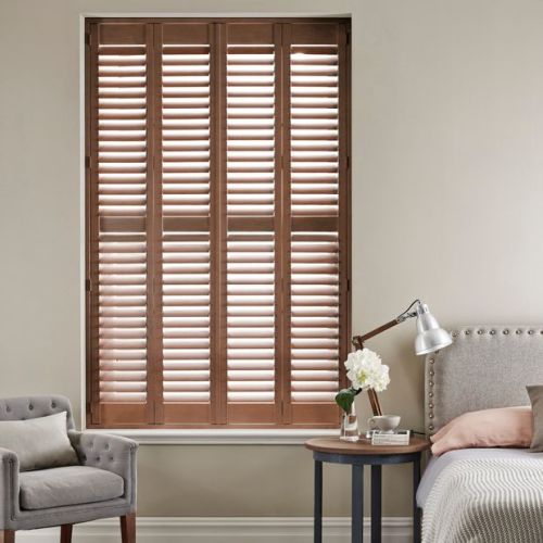 Wooden plantation shutters, four-panel and internal-mounted, closed with slats tilted to filter daylight; set in a bedroom corner with an armchair, nightstand, reading lamp, and neutral walls.