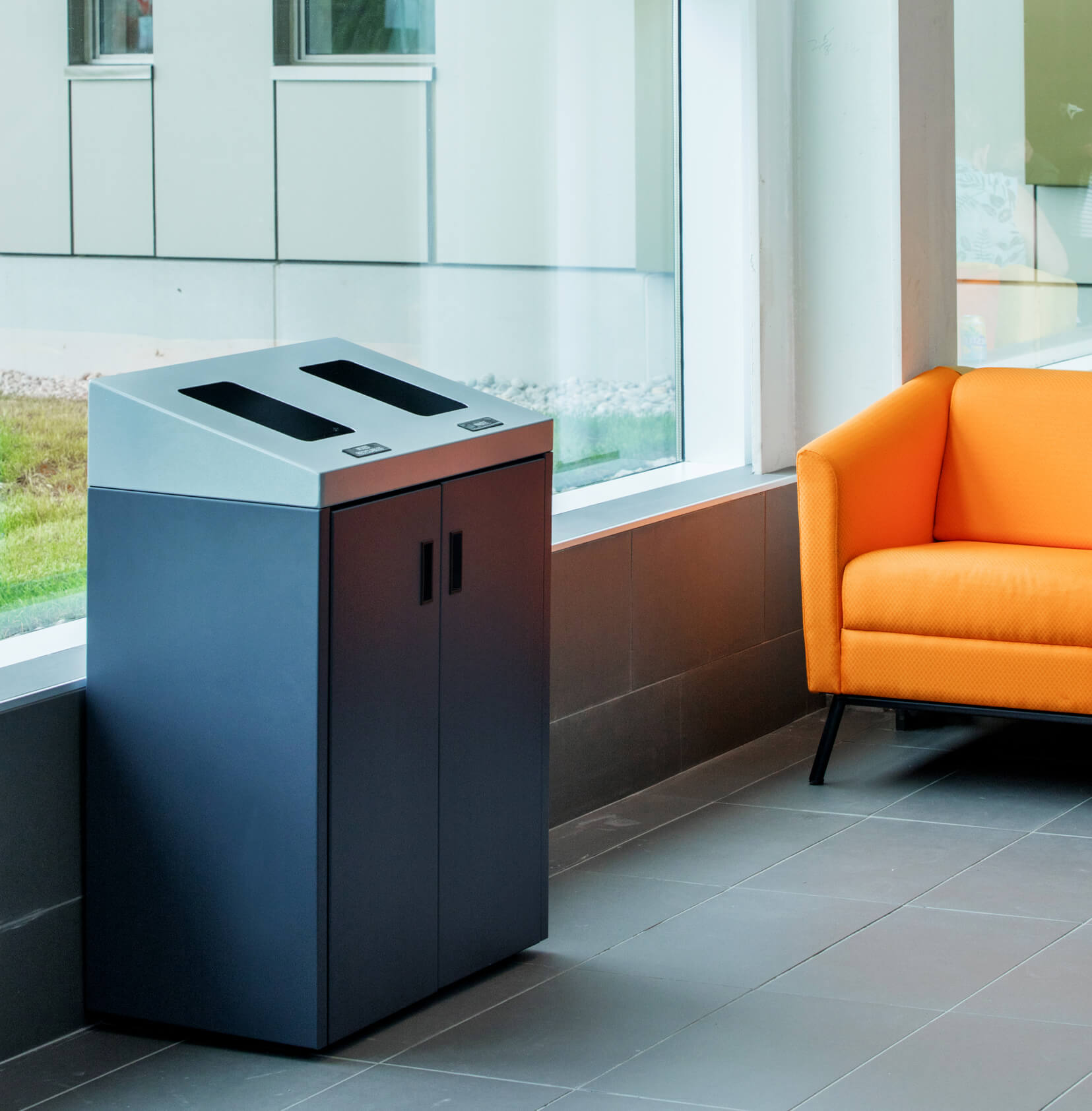waste and recycling bin with sloped lid and hinged front doors in a university campus beside an orange chair