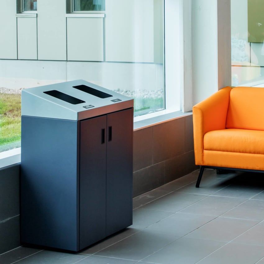 waste and recycling bin with sloped lid and hinged front doors in a university campus beside an orange chair