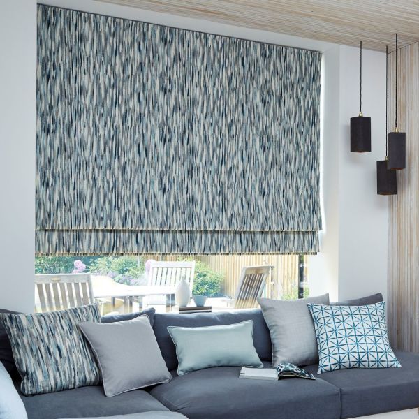 A blue-gray patterned Roman blind, lowered with a narrow bottom gap, filters daylight over a living-room window; grey sofa with cushions and pendant lights, patio garden visible beyond.