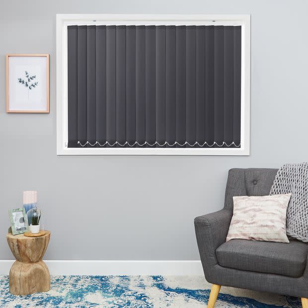 Dark grey vertical blinds fully closed, scalloped lower edges; blocking light in a grey-walled living room with a grey armchair, wooden stump side table, framed botanical print and blue rug.