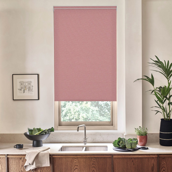 Rose-colored roller blind hangs nearly fully lowered over a kitchen window, filtering light; below stainless faucet and double sink sit on stone countertop with wooden cabinets, plants and bowls.