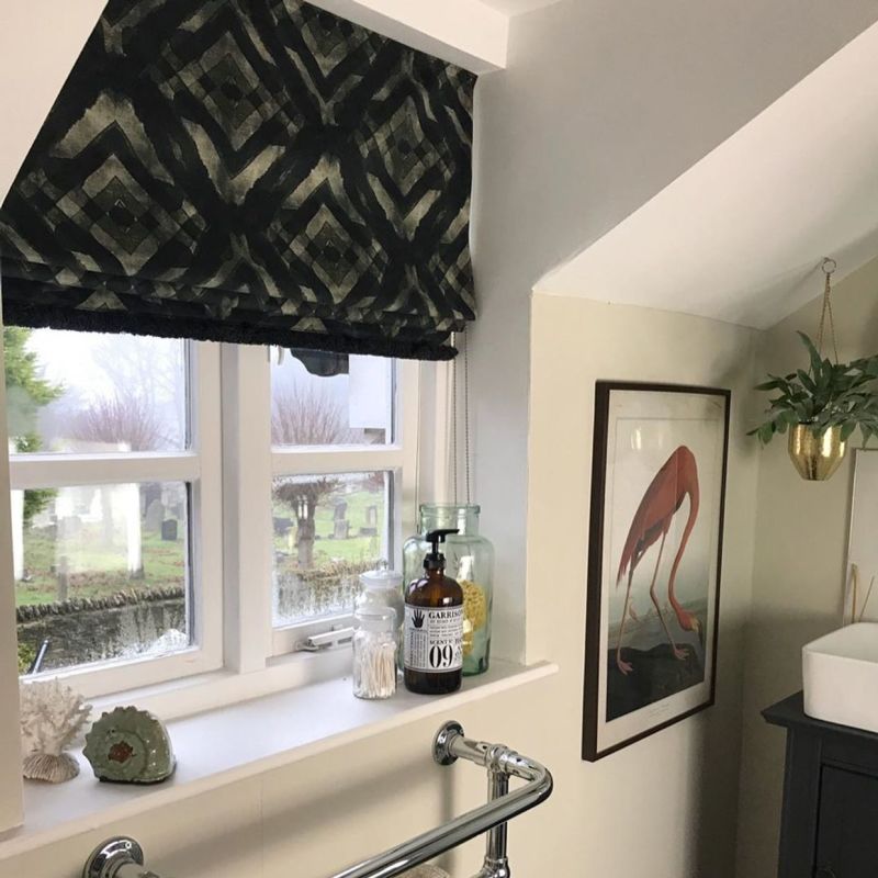Black-and-beige patterned Roman blind, partially lowered and dimming light; bathroom windowsill with soap dispenser and jars, framed flamingo print and hanging plant, misty graveyard visible outside.

I canâ€™t clearly read the label textâ€”please provide a close-up if you need exact transcription.