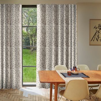White floor-length curtains with irregular brown spots hang across a tall, divided-pane window, partially drawn to reveal a green garden; mid-century dining table and chairs fill a sunlit room.