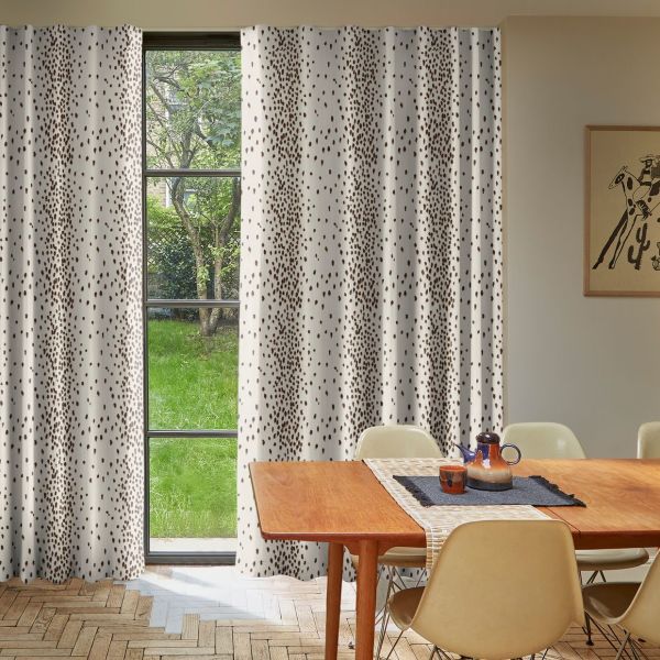 White floor-length curtains with irregular brown spots hang across a tall, divided-pane window, partially drawn to reveal a green garden; mid-century dining table and chairs fill a sunlit room.