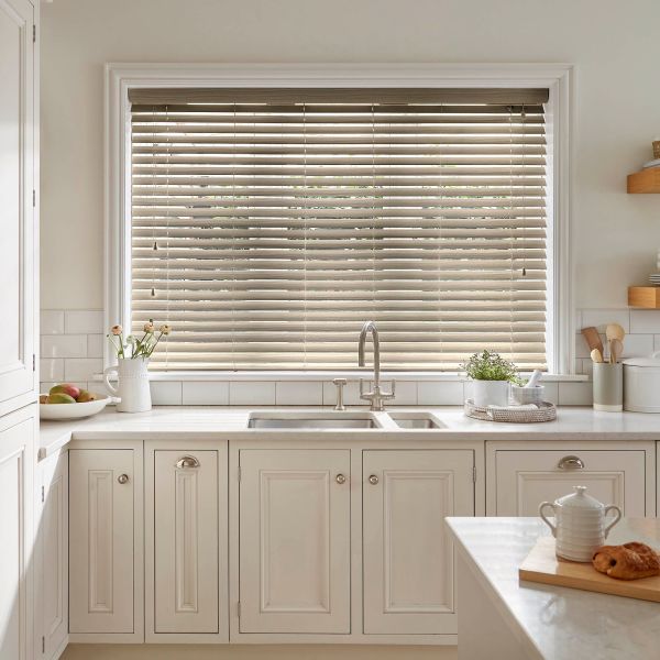Horizontal wooden blinds covering a large kitchen window, slats mostly closed filtering soft daylight; beneath, a stainless-steel sink with cream cabinetry and a marble countertop holding plants and kitchenware.