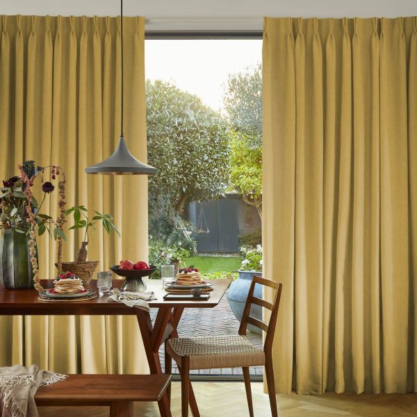 Floor-length pleated golden curtains, mostly drawn with a central opening, gently filtering daylight. They frame a dining table set for breakfast, pendant lamp overhead, and a lush backyard garden beyond.