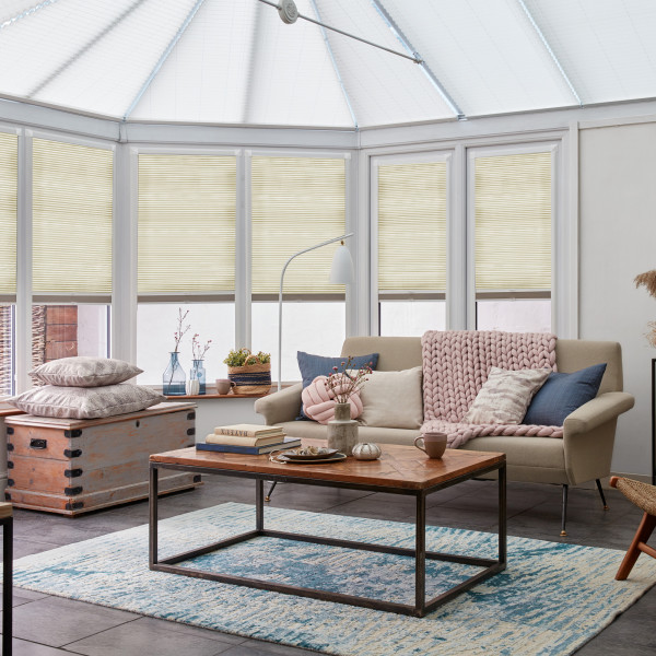 Beige pleated cellular blinds covering tall conservatory windows, lowered halfway and softly filtering daylight; a bright, cozy sunroom with a beige sofa, wooden coffee table, and blue patterned rug.