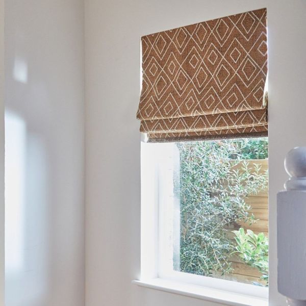 Fabric Roman blind, beige with white diamond pattern, partially lowered over a window, filtering daylight; small stair landing with walls and banister, outside view of shrubs and wooden fence.