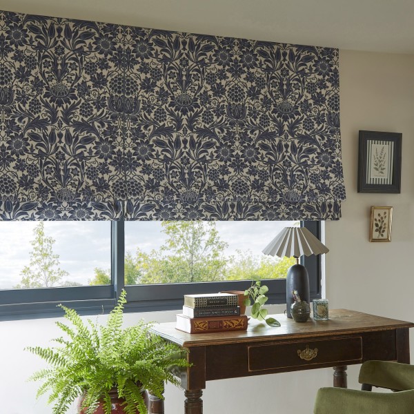 Patterned fabric Roman blind, lowered across the window with dense navy botanical print on cream, filtering daylight over a wooden desk with lamp, books, fern and a treed outside view.