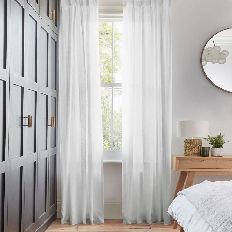 Sheer white pleated curtains, floor-length and hung on either side of a tall sash window, filtering soft daylight; bedroom setting with dark paneled wardrobe, wooden bedside table, lamp and mirror.