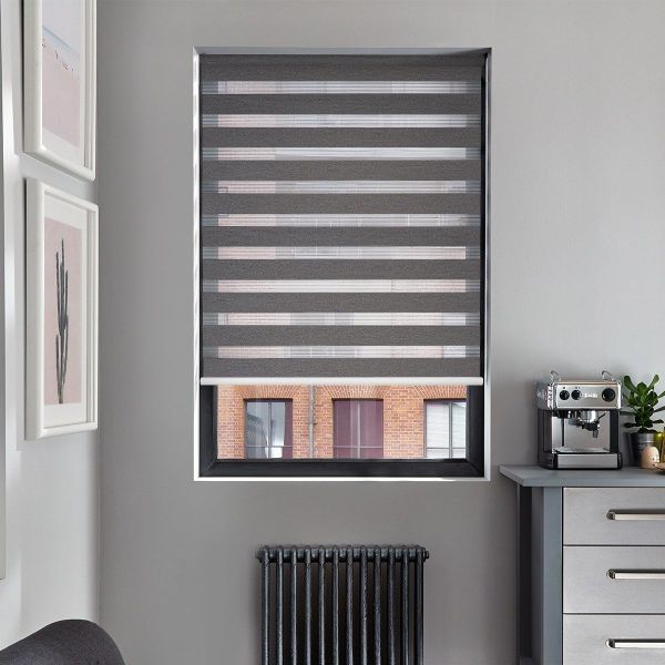 Grey zebra roller blind, lowered with alternating opaque and sheer horizontal stripes, filtering light and revealing a bottom view of a brick building; kitchen corner with radiator and espresso machine.