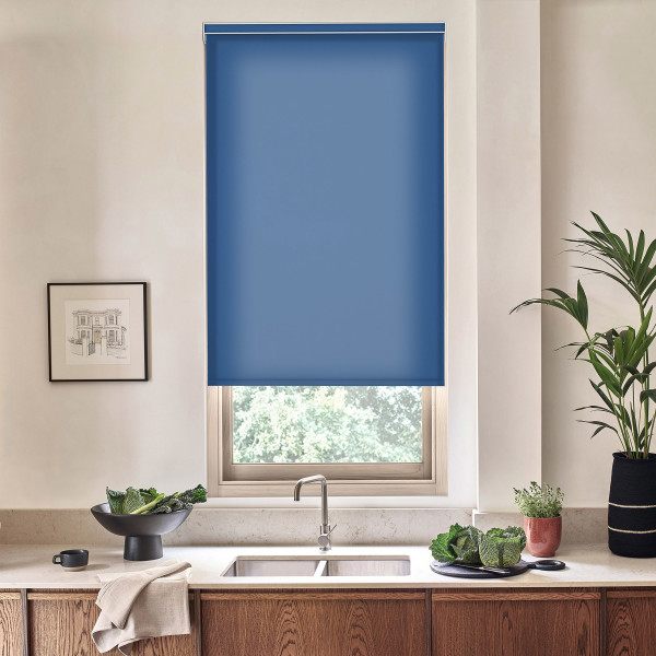 Blue roller blind covers most of a kitchen window, filtering daylight; below, a stainless faucet and sink sit on a marble countertop with bowls of leafy greens and potted plants.
