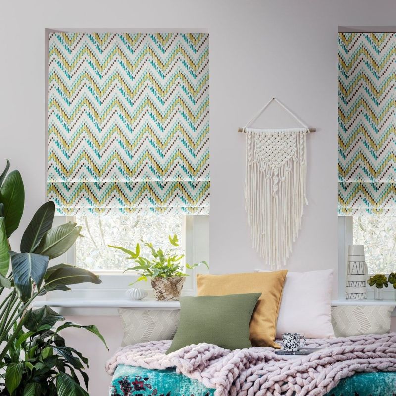 Chevron-patterned Roman shades cover the upper window panels, partially lowered and filtering daylight; a boho bedroom nook with macramÃ© wall hanging, potted plants, cushions, and a chunky knit throw.