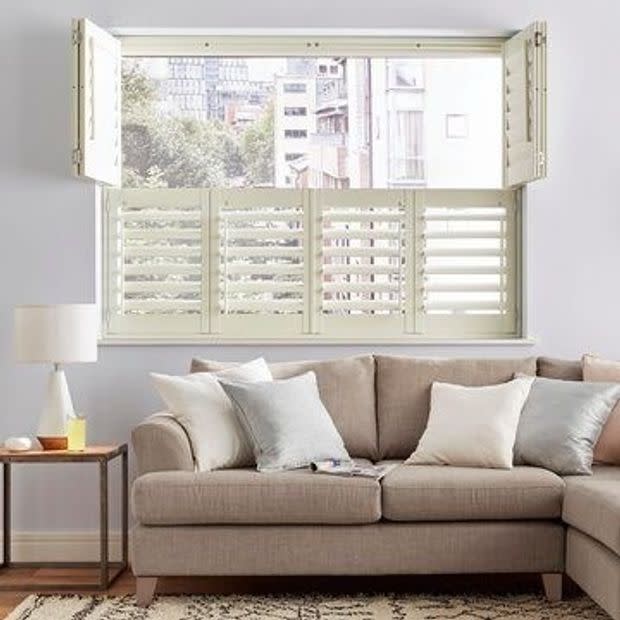 Cream louvered plantation shutters across lower sash, top hinged shutters folded outward; louvers partially open filtering daylight, city view; beige sofa with pillows, side table and lamp in living room.