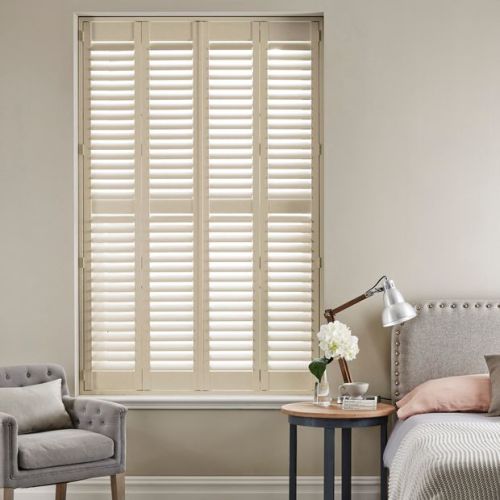 Cream plantation shutters fill a recessed window, full-height panels with horizontal louvers angled to admit light, filtering bright daylight into a neutral bedroom with armchair, bedside table and lamp.