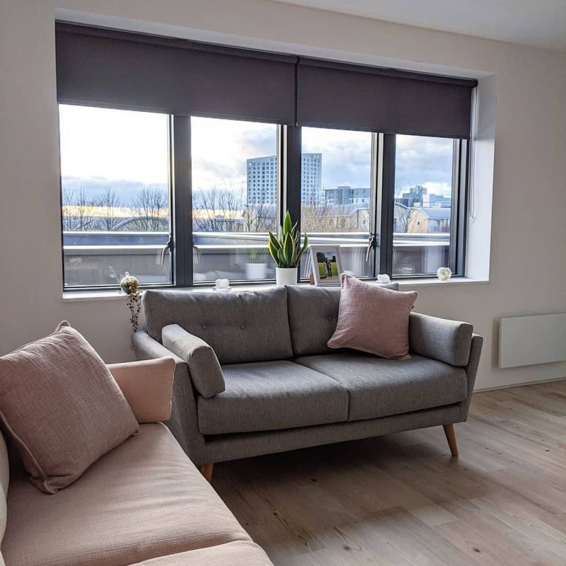 Dark grey roller blinds across a three-panel bay window, partially lowered and blocking light; overlooking a cloudy cityscape. Living room with grey sofa, pink cushions, wooden floor and plant.