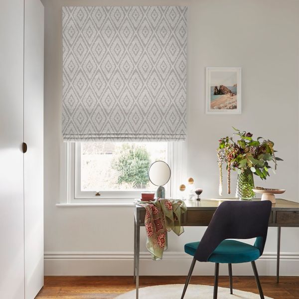 Grey geometric diamond-patterned Roman blind, lowered over the window and filtering soft daylight; small home office desk beneath with teal chair, vase of flowers, makeup mirror and framed coastal print.