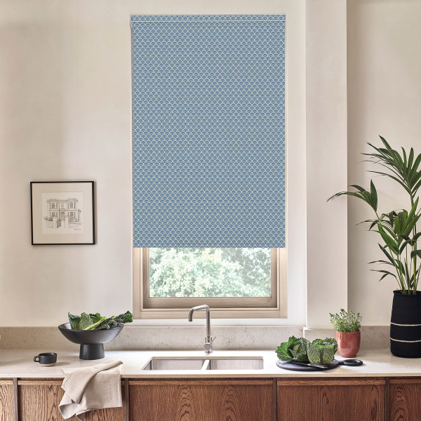 A blue patterned roller blind covers a window above a kitchen sink, partially lowered; the countertop holds a bowl of leafy greens, potted plants, a faucet, and a framed sketch on the wall.