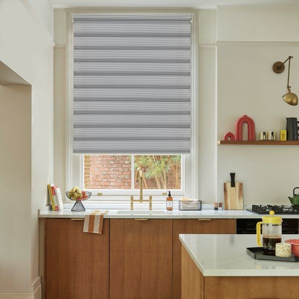 Gray cellular roller blind with horizontal striped bands, lowered to cover most of the window and filtering daylight, above a kitchen sink with brass faucet, marble countertop and wooden cabinets.