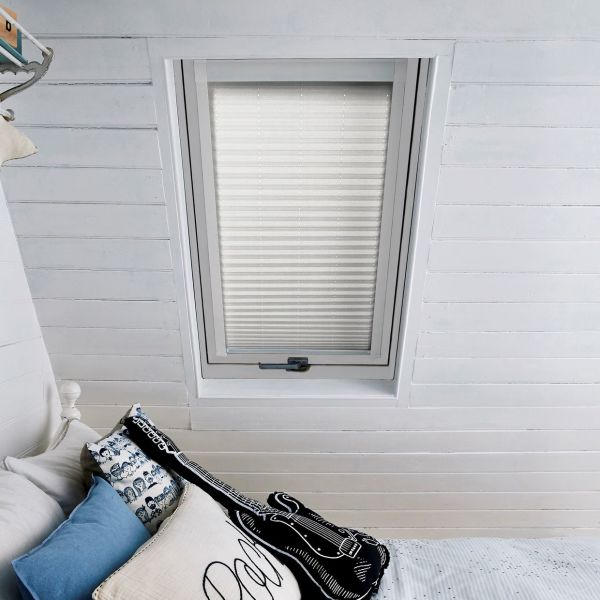 White pleated blind, fully lowered in a skylight recess with fine horizontal folds; diffusing daylight in a white-painted slatted attic bedroom above a bed with pillows and a guitar cushion.