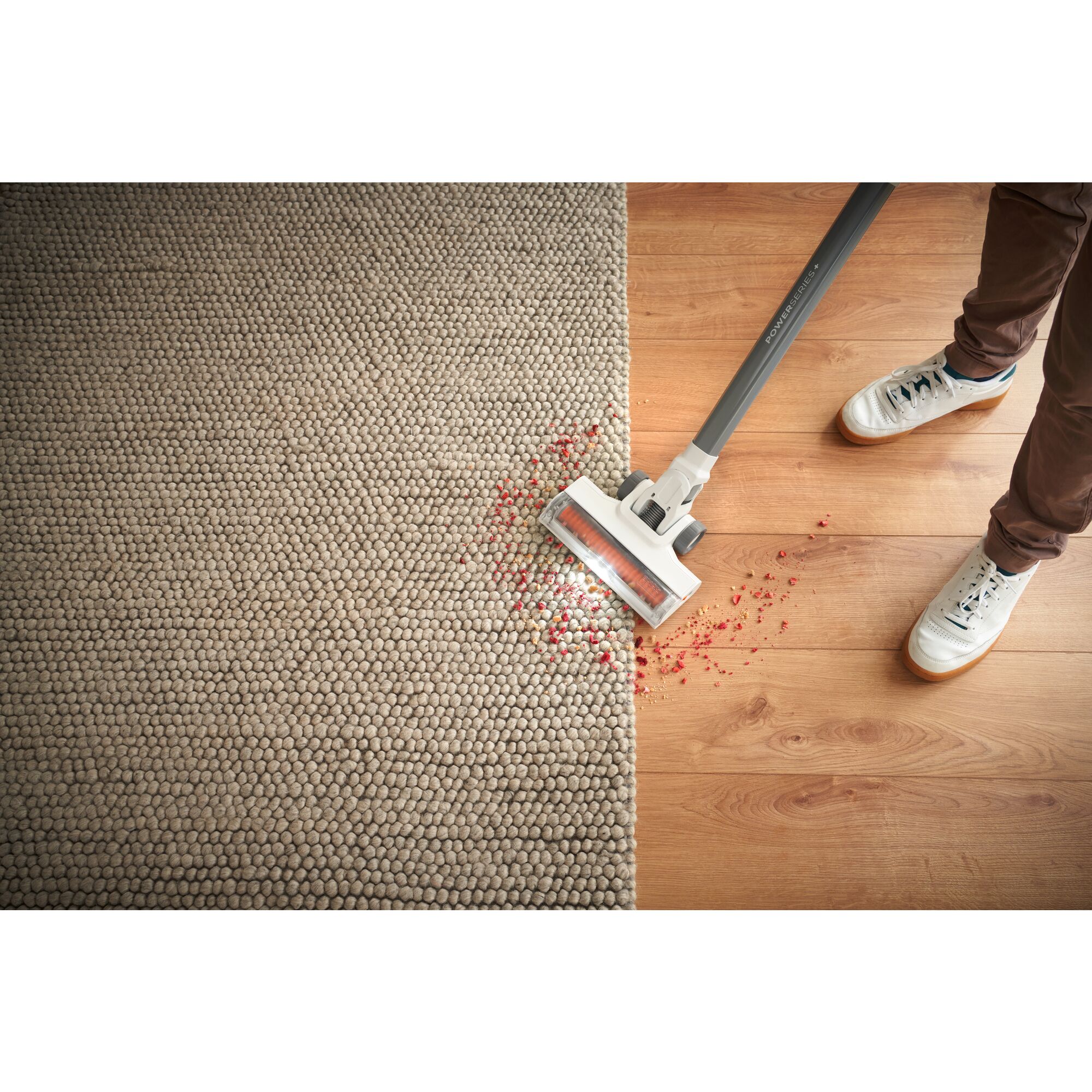POWER SERIES cordless stick vacuum being used to clean debris on wooden floor and rug.