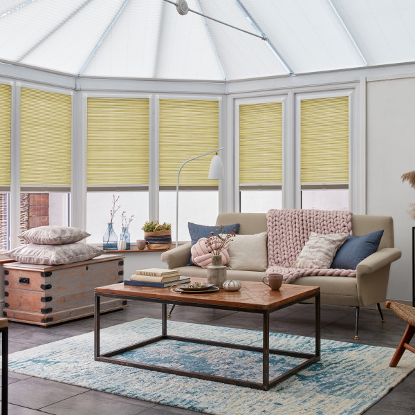 Honey-yellow pleated blinds, lowered across six tall conservatory windows, filtering soft daylight; beige sofa with knitted throw faces a wooden coffee table on a blue-pattern rug in a bright sunroom.