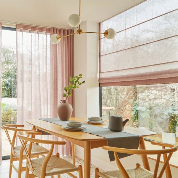 Sheer dusty-pink floor-to-ceiling curtains at left and a lowered matching Roman blind on the right, gently filtering soft daylight over a wooden dining table with chairs and garden view.
