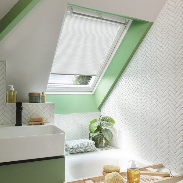 White pleated skylight blind, mostly lowered over a roof window, filtering daylight; bathroom with green-painted sloped ceiling, white herringbone tiles, a sink and tub, plant, and toiletries.