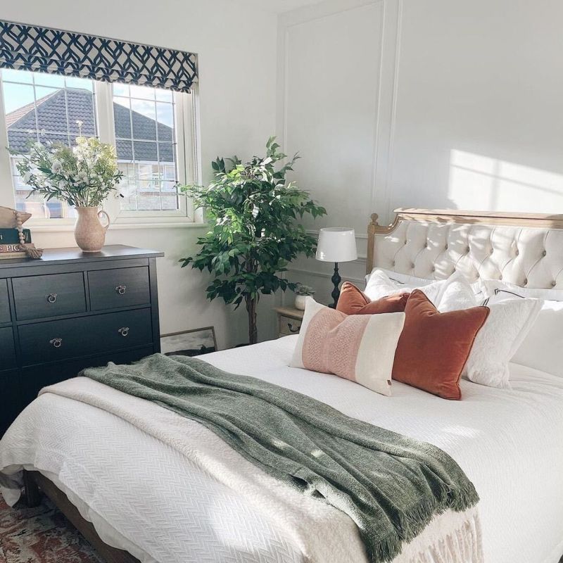 Navy geometric Roman valance mounted above a mullioned window, filtering soft morning light; bedroom setting with tufted headboard, white bedding, rust cushions, green throw, dresser and potted plant.