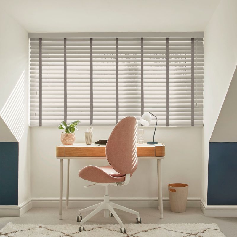 Horizontal slatted blinds over a wide window, pale grey with tapes and cord, closed and filtering daylight, casting striped shadows across a study with wooden desk and pink swivel chair.