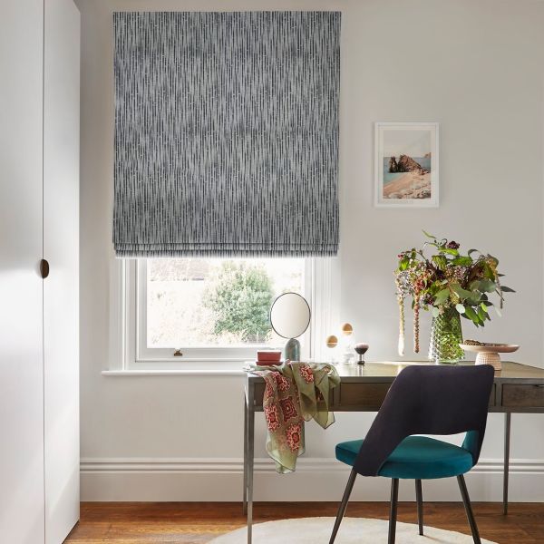 Gray patterned Roman blind lowered over window, filtering daylight; wooden desk with round mirror, vase of flowers and scarf beside a teal chair in a lit office with garden view.