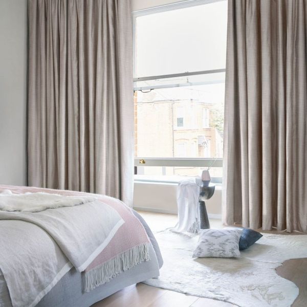 Floor-length beige linen curtains, gathered at both sides and partially covering a tall sash window, filtering soft daylight into a neutral bedroom with a bed, rug, and street view.