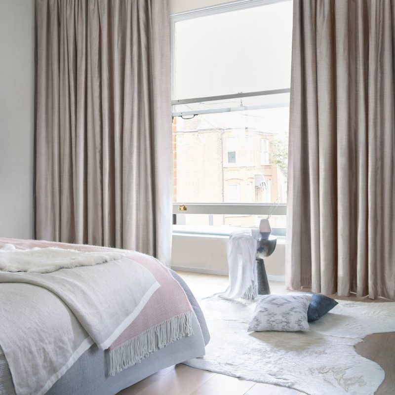 Floor-length beige linen curtains, gathered at both sides and partially covering a tall sash window, filtering soft daylight into a neutral bedroom with a bed, rug, and street view.