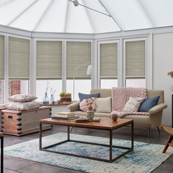 Pleated cellular blinds covering five tall bay windows, lowered to mid-height and filtering soft daylight; bright conservatory-style living room with beige sofa, wooden coffee table and pastel accents.