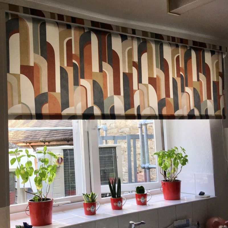 Retro-arched patterned roller blind, lowered across the window, filtering light; kitchen windowsill beneath holds red potted herbs and small succulents, outside view of a stone wall and metal pipes.