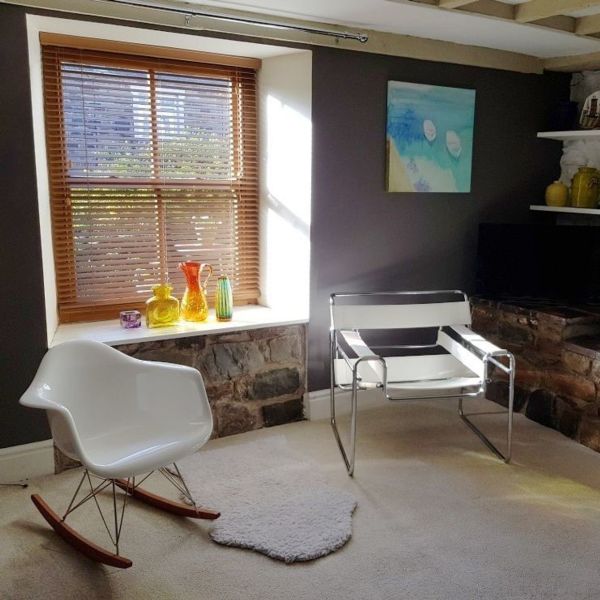 Wooden horizontal blinds in recessed window, partially closed, filtering sunlight onto a sill with colorful glass vases; living-room corner with stone ledge, two modern chairs and gray walls.