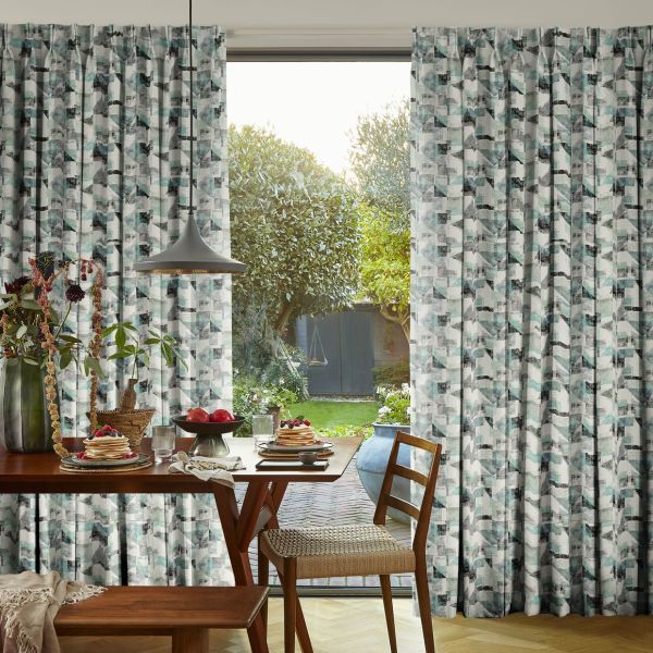 Pleated teal-gray geometric curtains, full-length, frame a central open sliding door, parted to admit sunlight; a dining table set for breakfast overlooks a garden with trees, swing, and shed.