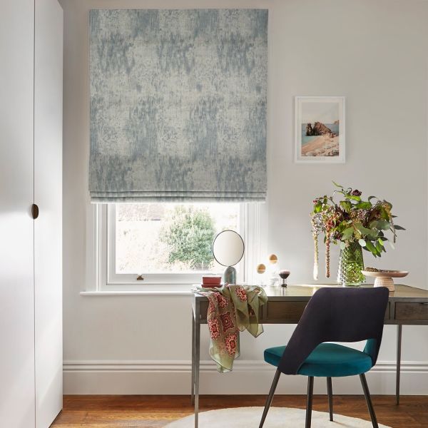 A textured blue-gray Roman blind hangs fully lowered, softly filtering daylight over a desk; room includes a window-facing wooden desk with vase of flowers, teal chair, and framed seaside print.
