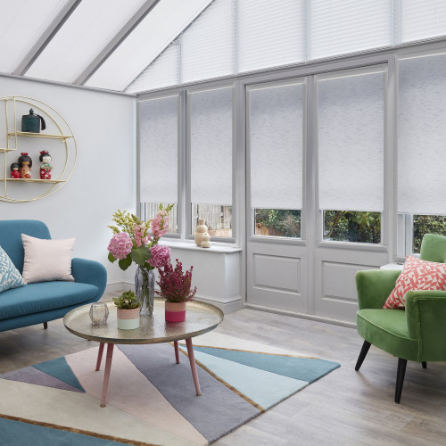 White roller blinds and pleated roof blinds covering tall glass doors and windows, partially lowered and filtering soft daylight in a bright conservatory with colorful seating, geometric rug and plants.