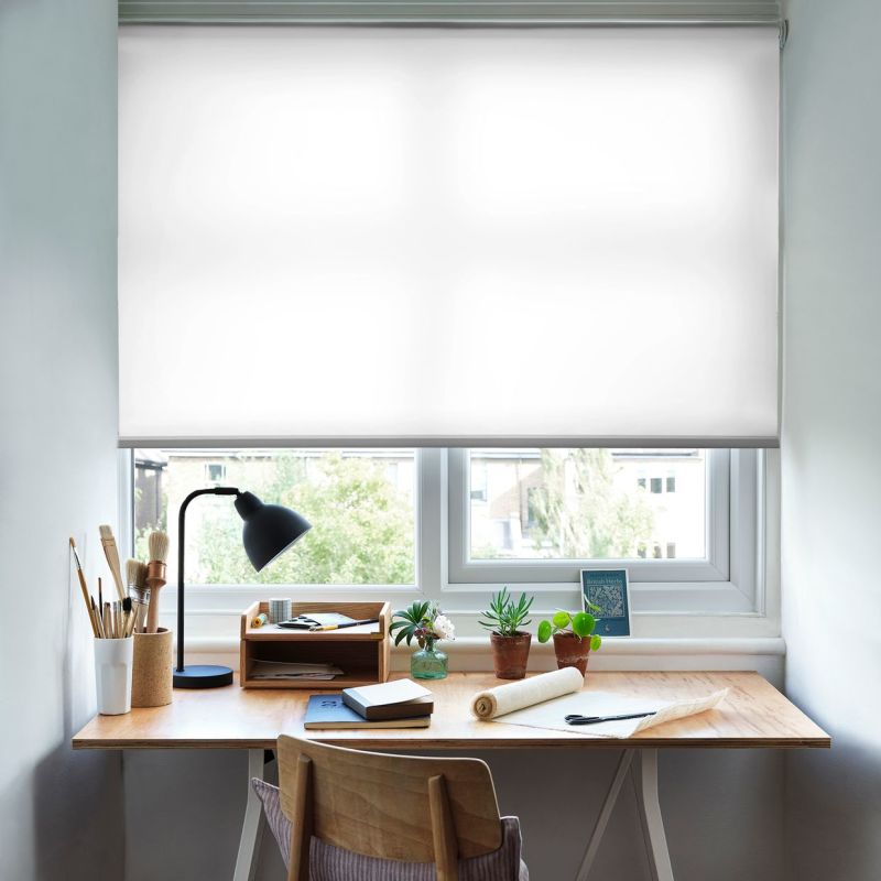 White roller blind, lowered nearly fully, translucent fabric softly filtering daylight; above a wooden desk in a bright home workspace featuring a lamp, art brushes, notebooks and small potted plants.