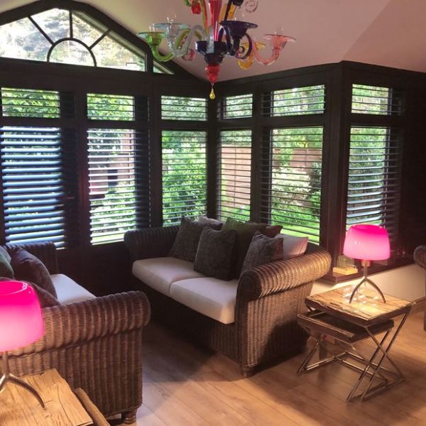 Dark wooden plantation shutters cover a bay of tall windows, slats partially open, filtering garden light into a cozy sunroom with wicker sofas, neutral cushions, wooden floor and colorful chandelier.