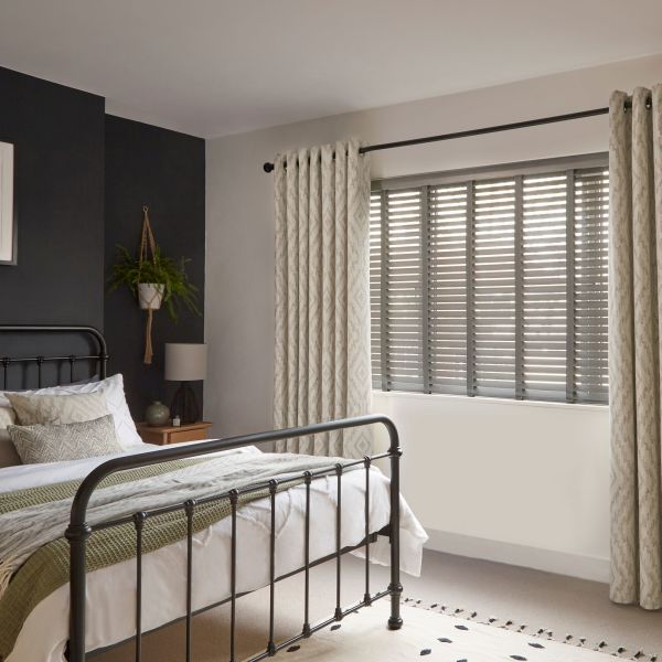 Patterned grommet curtains flank a window with horizontal slatted blinds; curtains pulled aside, blinds tilted to filter daylight in a softly lit bedroom with a black metal bedframe.