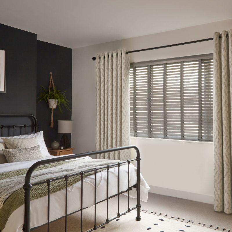 Patterned grommet curtains flank a window with horizontal slatted blinds; curtains pulled aside, blinds tilted to filter daylight in a softly lit bedroom with a black metal bedframe.
