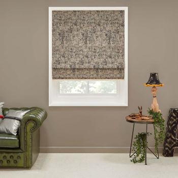Textured beige-gray Roman shade, mostly lowered with visible hem, filtering daylight through a white-framed window; set in a neutral living room with green leather sofa, side table, lamp, and trailing plant.