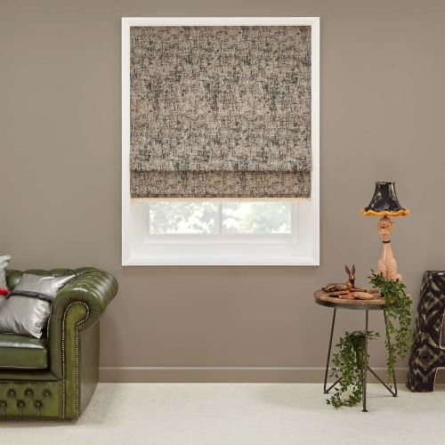 Textured beige-gray Roman shade, mostly lowered with visible hem, filtering daylight through a white-framed window; set in a neutral living room with green leather sofa, side table, lamp, and trailing plant.