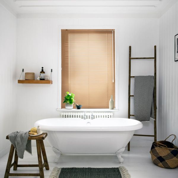 Horizontal wooden Venetian blinds covering a central window, fully lowered and closed, filtering warm daylight; bathroom with white clawfoot tub, wooden stool, ladder towel rack, and potted plant on the sill.