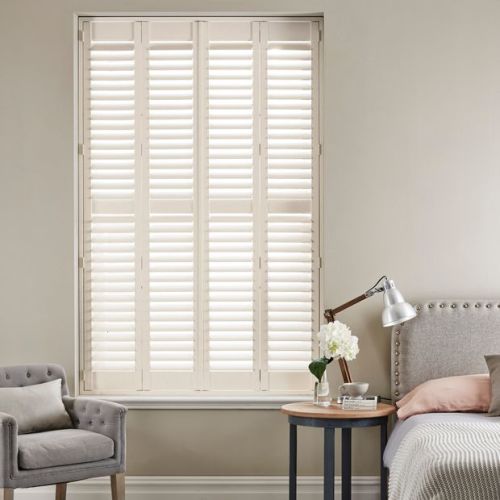 White wooden plantation shutters covering a tall window, louvered panels closed but allowing diffused light; set in a neutral-toned bedroom with armchair, bedside table, lamp, and flowers.